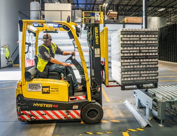 A warehouse worker maneuvers a forklift to transport crates for brewing company storage.